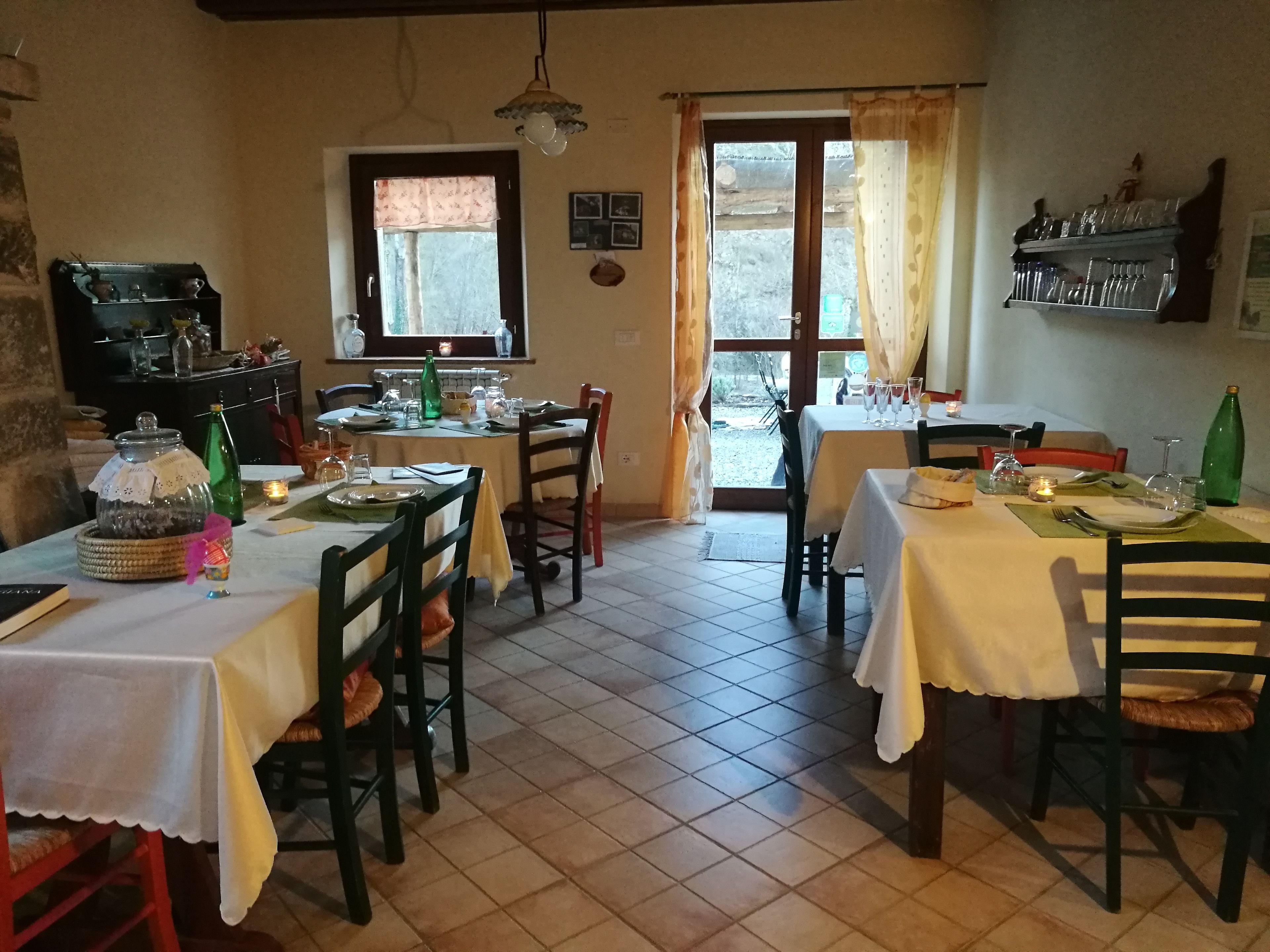 A dining area with two tables set for meals, chairs, a window with curtains, a shelf with bottles, and a tiled floor.