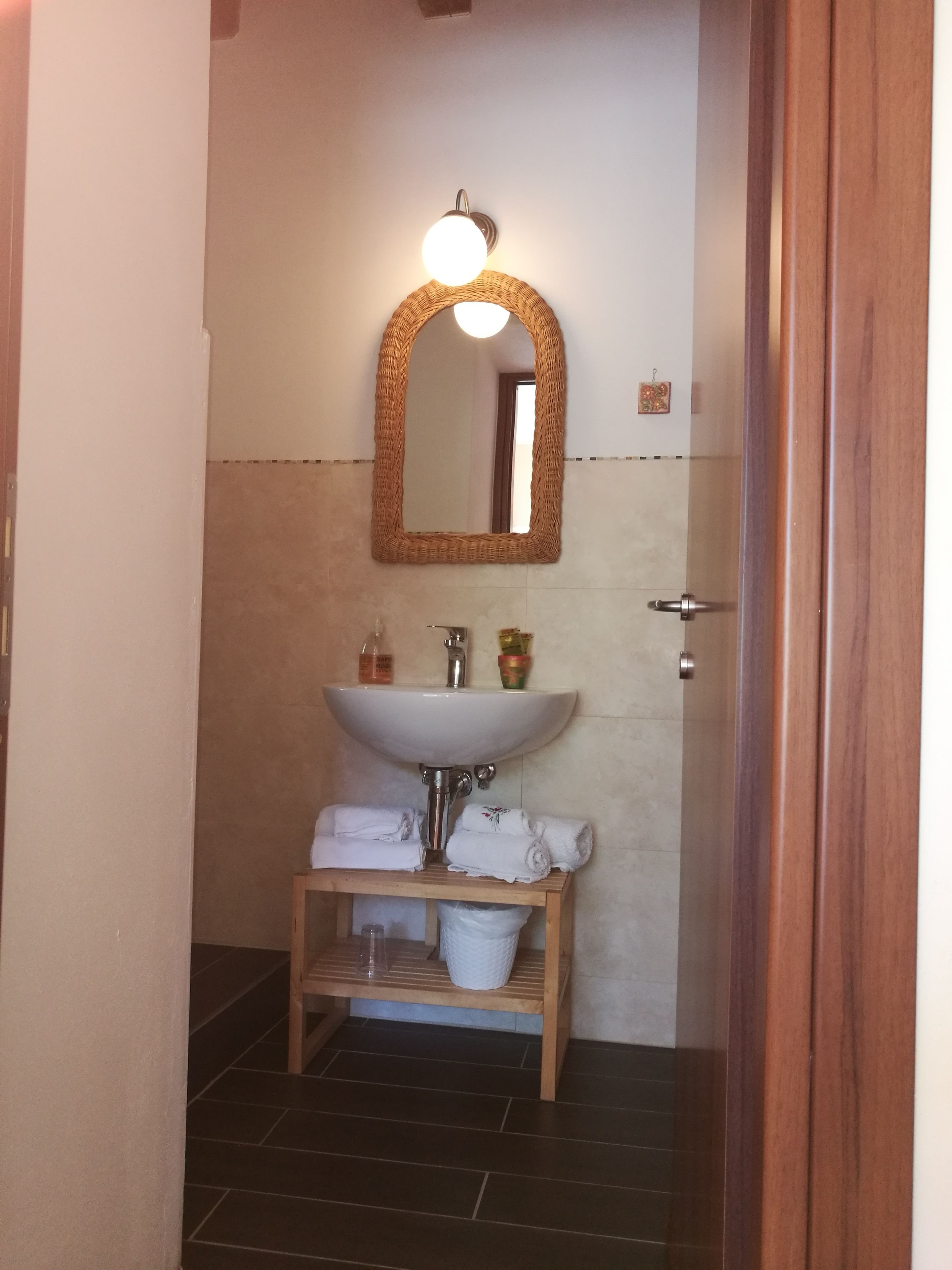 Une salle de bain avec un lavabo blanc, un miroir avec un cadre en osier et une étagère en bois où sont posées des serviettes.