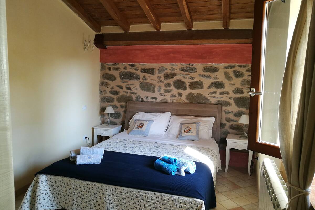 Une chambre avec un mur en pierre, des poutres en bois au plafond, un lit recouvert d’une couverture bleue et une table de chevet avec une lampe.