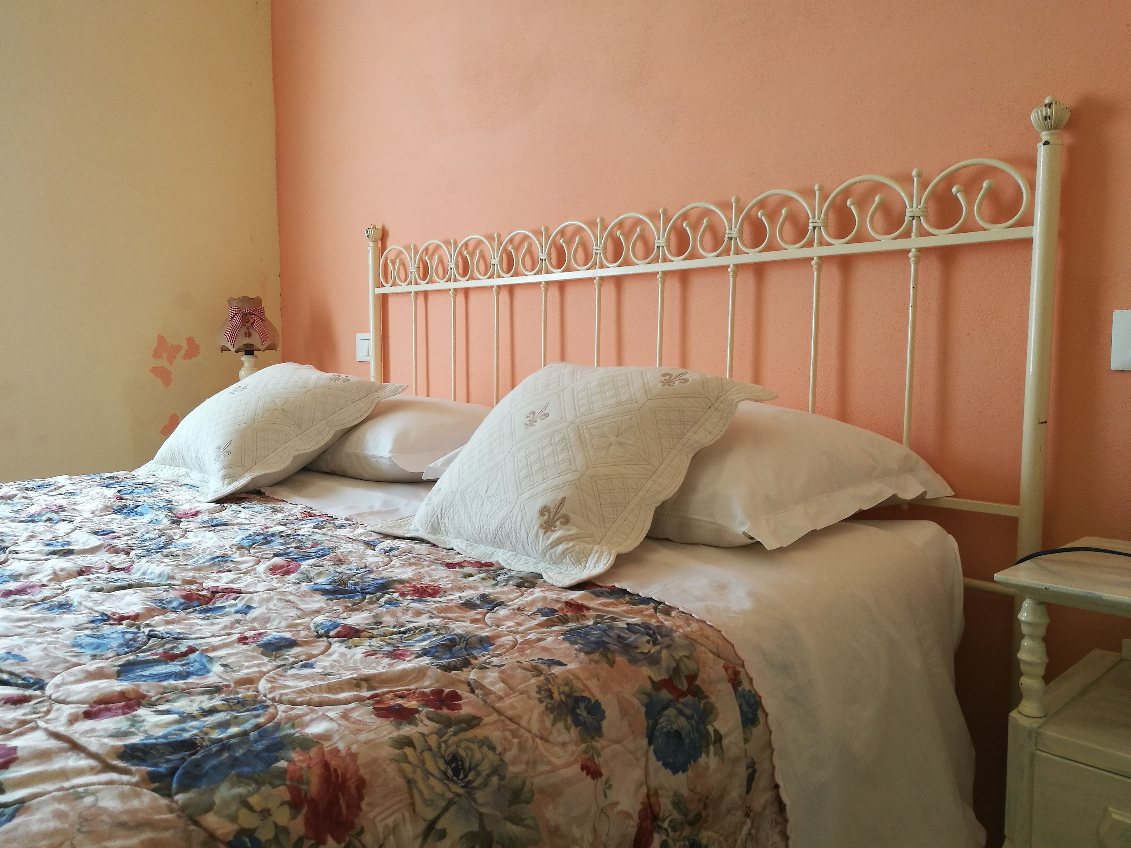 Una cama con colcha estampada con motivos florales y almohadas blancas, cabecero de metal blanco y mesita de noche con lámpara.