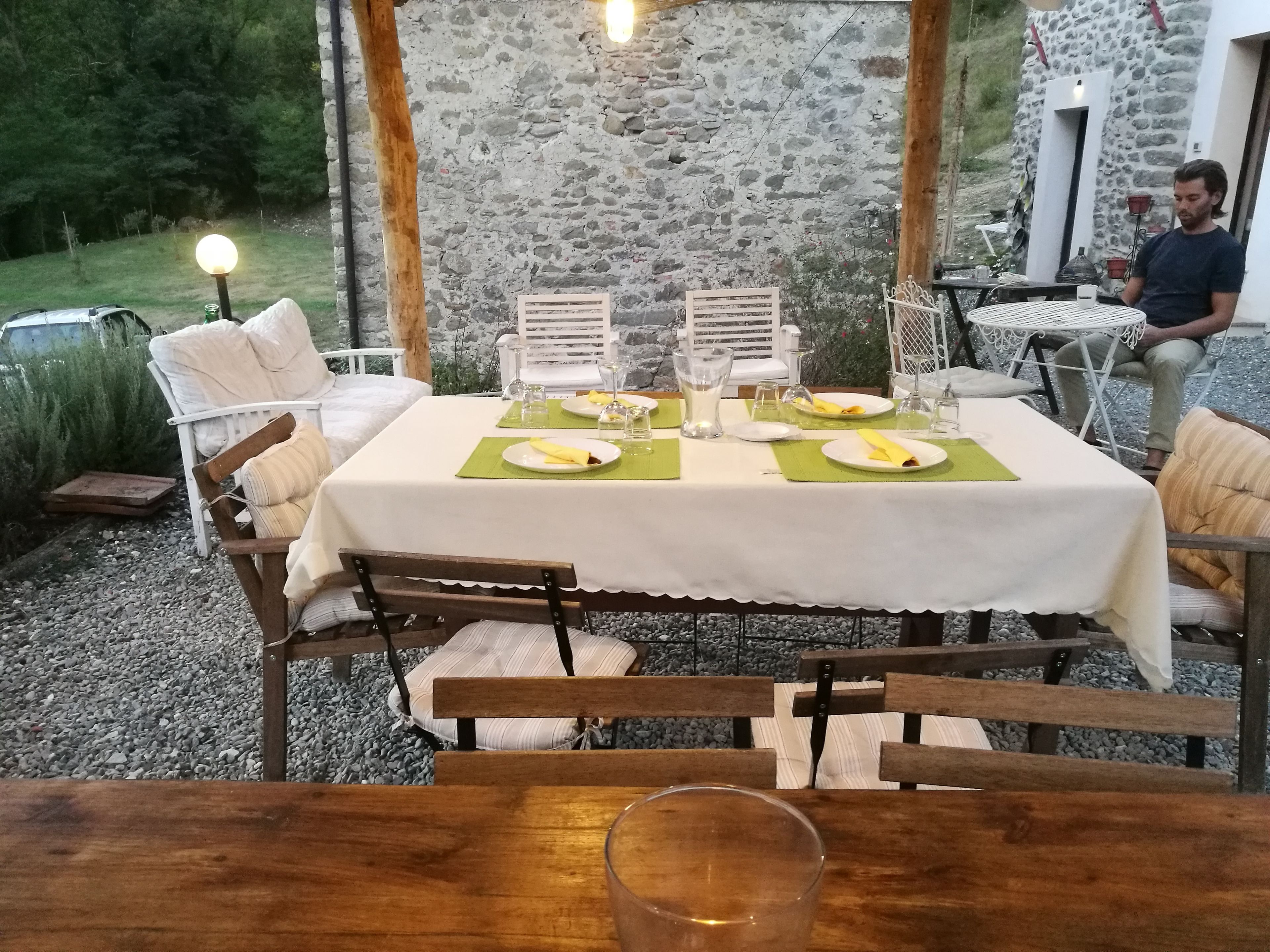 An outdoor dining area with a stone wall backdrop, a table set with plates, glasses, and cutlery, and a person seated at a table in the background.