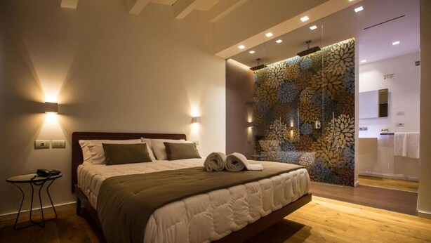 Ein modernes Hotelzimmer mit einem großen Bett, einem kleinen Nachttisch und einem durch eine Glaswand abgetrennten Badezimmer.