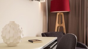 Ein modernes Zimmer mit einem weißen Tisch, einer geometrischen Vase, einem Buch, einer roten Lampe und einem hölzernen Hocker.