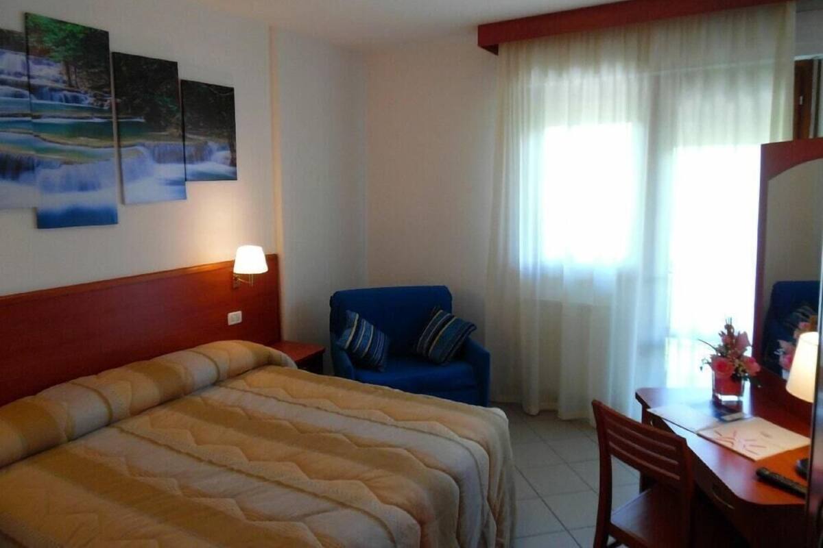 Une chambre d’hôtel avec un lit, un bureau, une chaise, une lampe et une fenêtre avec des rideaux.