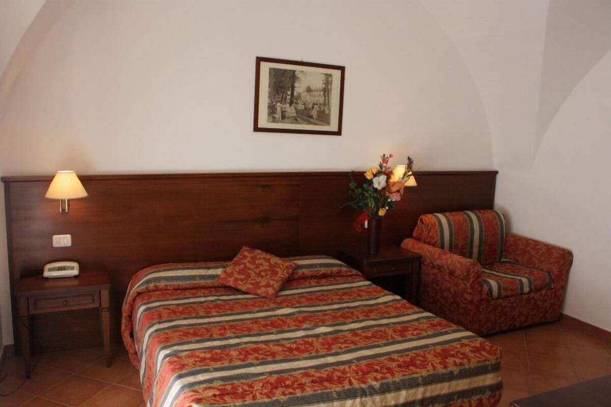 Une chambre d’hôtel avec une tête de lit en bois, un lit avec une literie à rayures, une table de chevet avec une lampe, une chaise et un tableau encadré au mur.