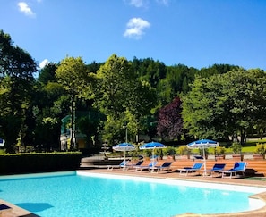 Una piscina con sillas reclinables azules y sombrillas, rodeada de árboles y un cielo despejado.