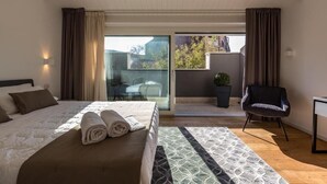A modern hotel room with a large bed, a small desk, a chair, a balcony with a view of buildings and trees, and a potted plant. - Junior Suite with Terrace - A wisp of smoke (Catania)