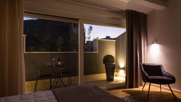 A hotel room with a bed, a small table with chairs, a potted plant, and a view through a sliding door to the outside. - Junior Suite with Terrace - A wisp of smoke (Catania)