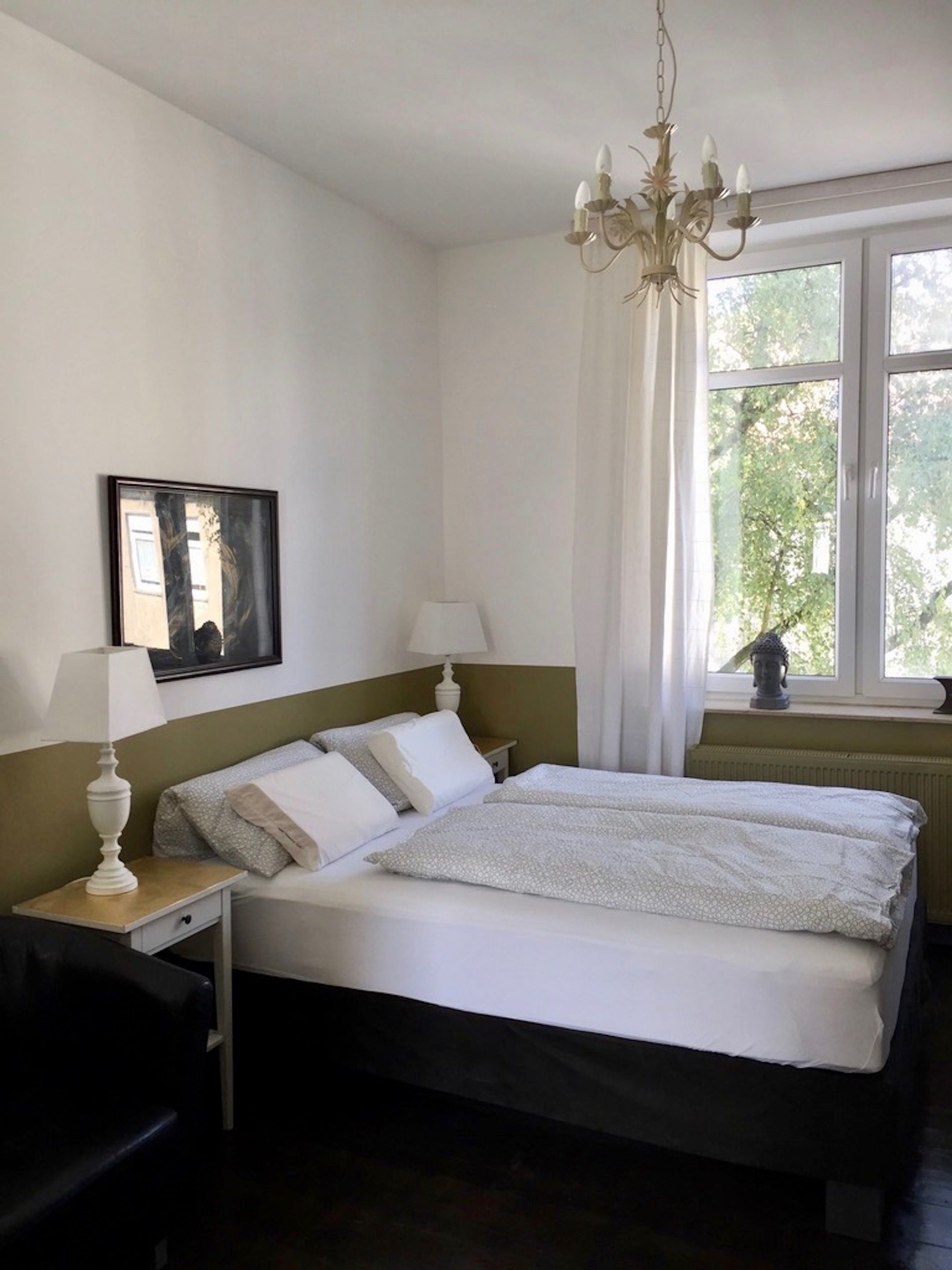 Ein ordentlich eingerichtetes Schlafzimmer mit Bett, Nachttischlampen, einem Nachttisch, einem Sessel, einem Fenster mit Vorhängen und einer Deckenleuchte.