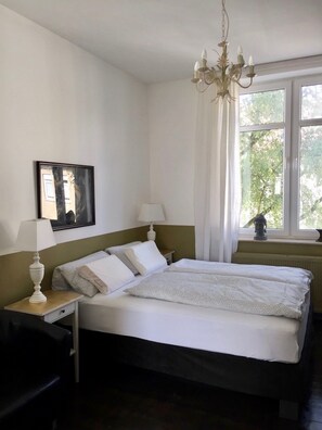 A neatly arranged bedroom with a bed, bedside lamps, a nightstand, a chair, a window with curtains, and a chandelier. - a-domo Apartments Oberhausen - Studio Apartments & Flats - short or longterm - single or grouptravel (Oberhausen)