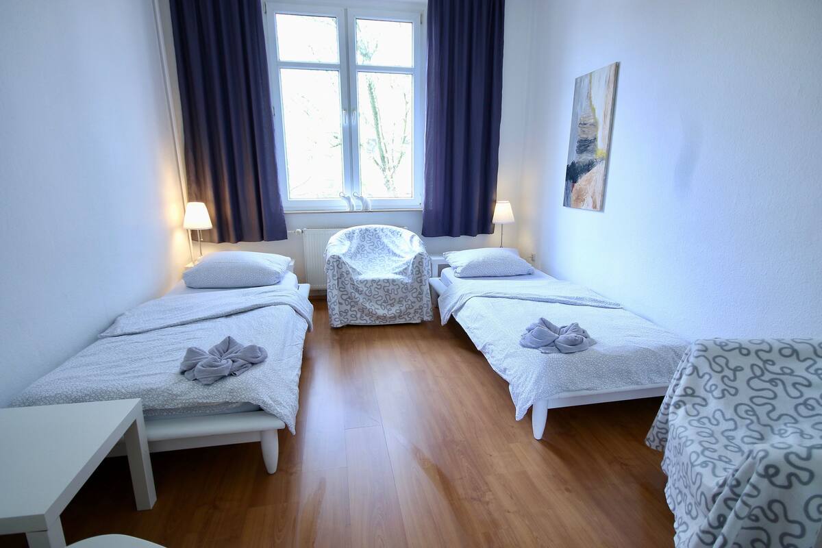 A room with two single beds, a small table, a chair, and a radiator.