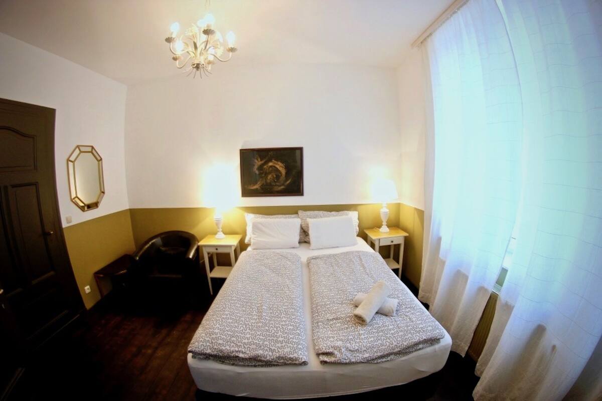 A hotel room with a large bed, two bedside tables, a chandelier, a framed artwork on the wall, and a door.