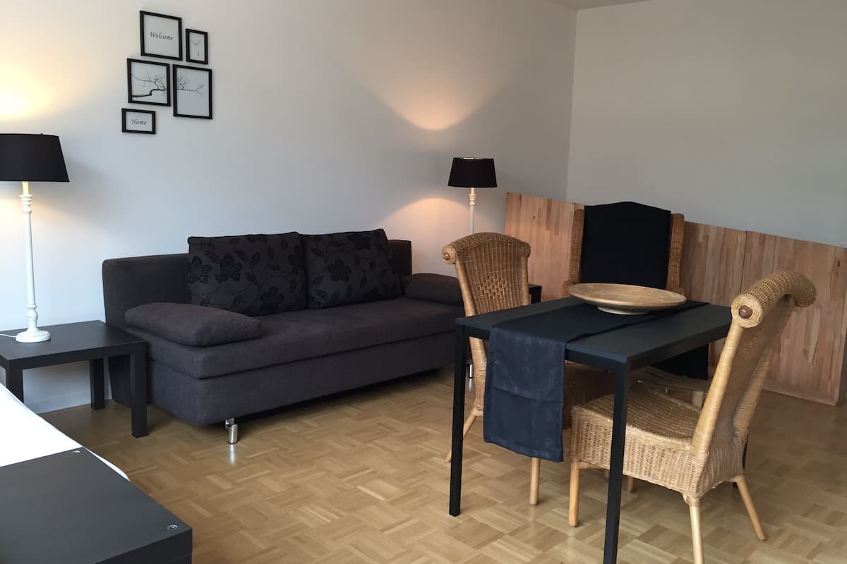 A modern living room with a sofa, a dining table, and two chairs. There are wall-mounted picture frames and two floor lamps.