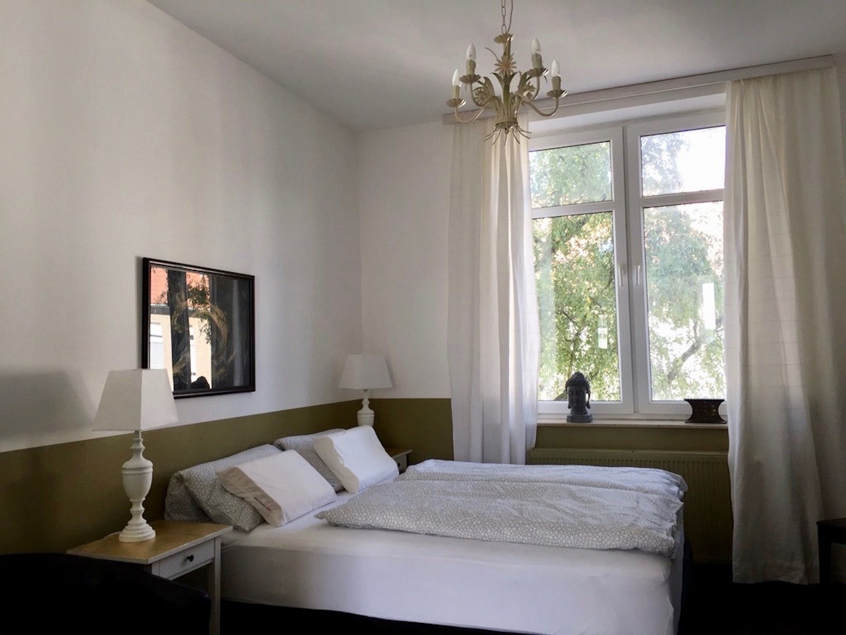 Ein Schlafzimmer mit Bett, Nachttisch, Lampe, einem Bild an der Wand und einem Fenster mit Vorhängen.