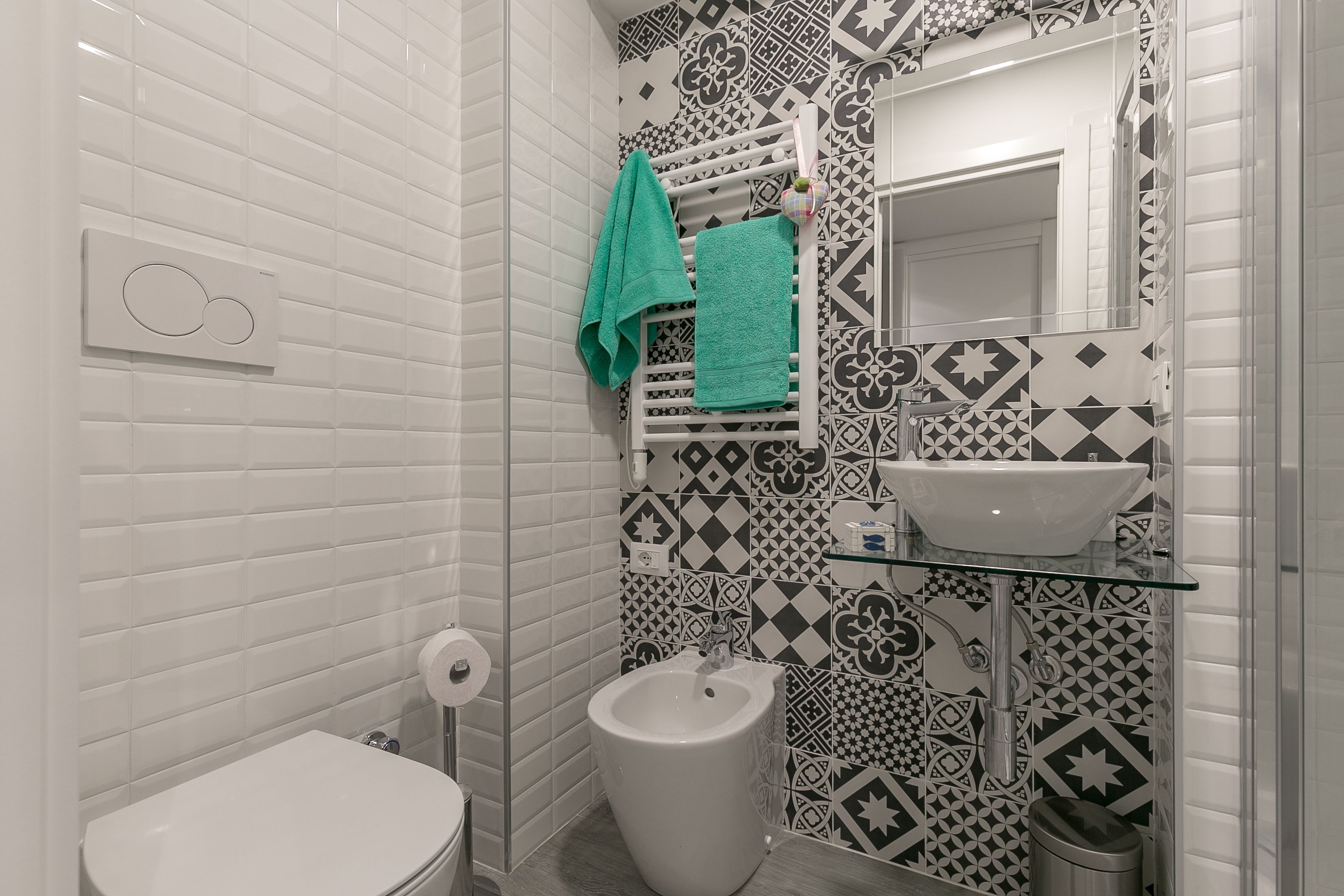 A bathroom with white tiled walls, a bidet, a sink with a glass shelf, and a patterned backsplash.
