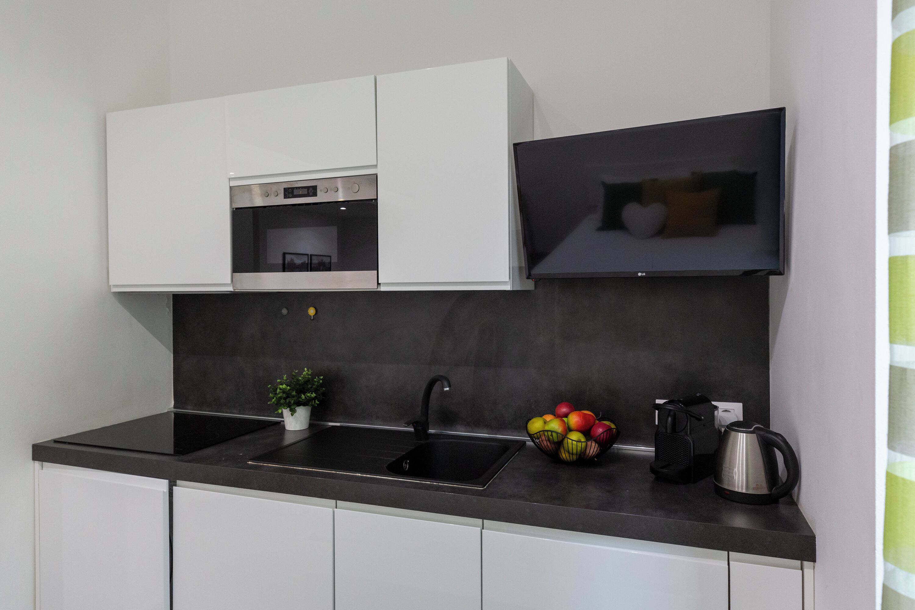 Una cucina moderna con mobili bianchi, un piano di lavoro nero, forno integrato, lavello nero, una pianta in vaso, una ciotola di frutta e un bollitore.