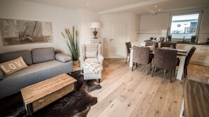 Un salon moderne avec un canapé gris, une table basse en bois et un tapis en peau de vache. On peut également apercevoir un fauteuil blanc et une table à manger avec des chaises.