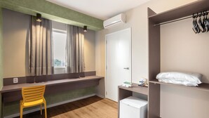 Een compacte hotelkamer met een houten vloer, een klein bureau met stoel, een garderobe en een minibar.