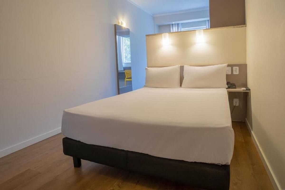 A neatly made bed in a room with a wooden floor, a bedside table with a lamp, and a window with curtains.