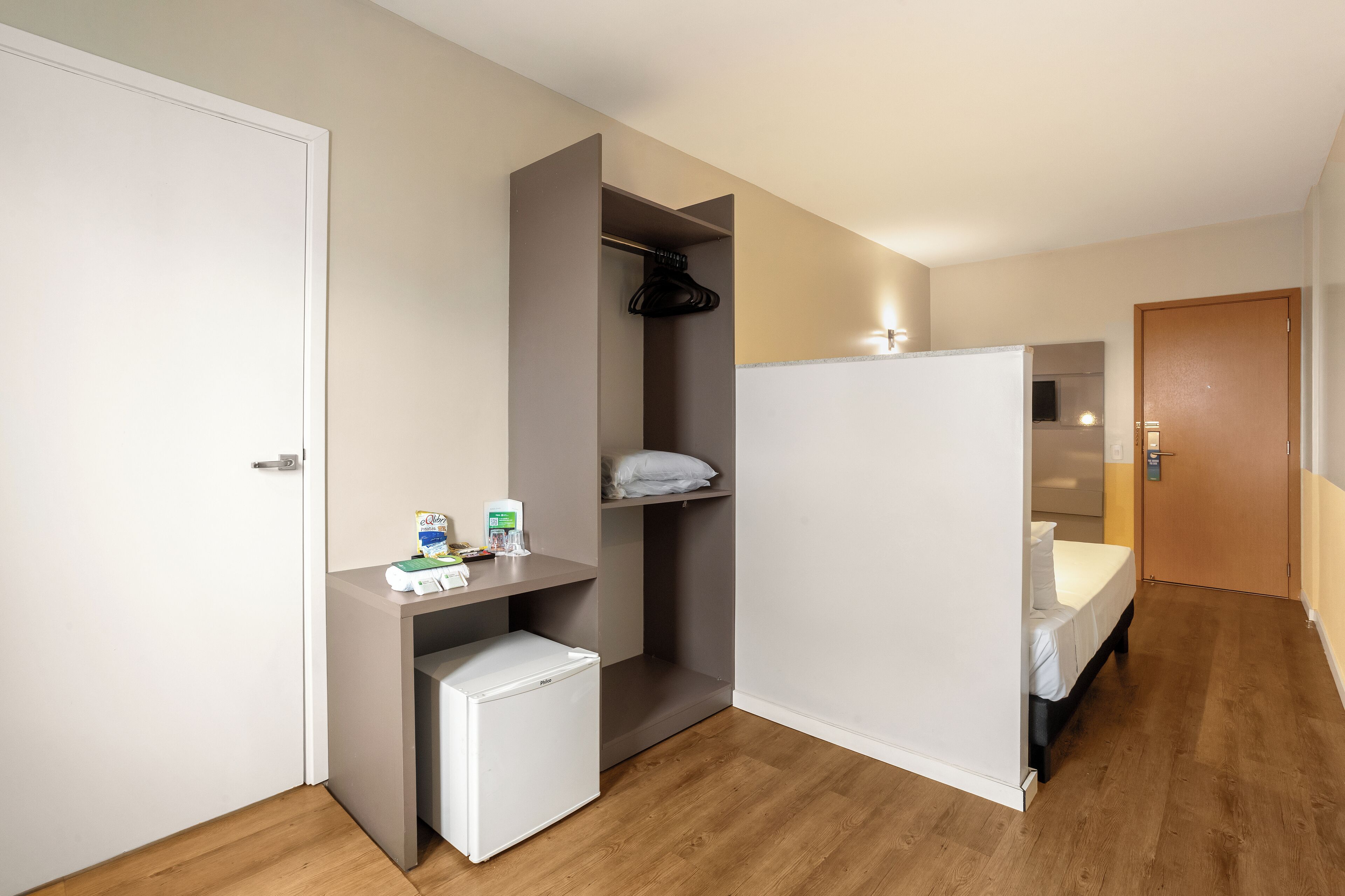 A compact room with a bed, a small refrigerator, a desk with a chair, and a wardrobe.