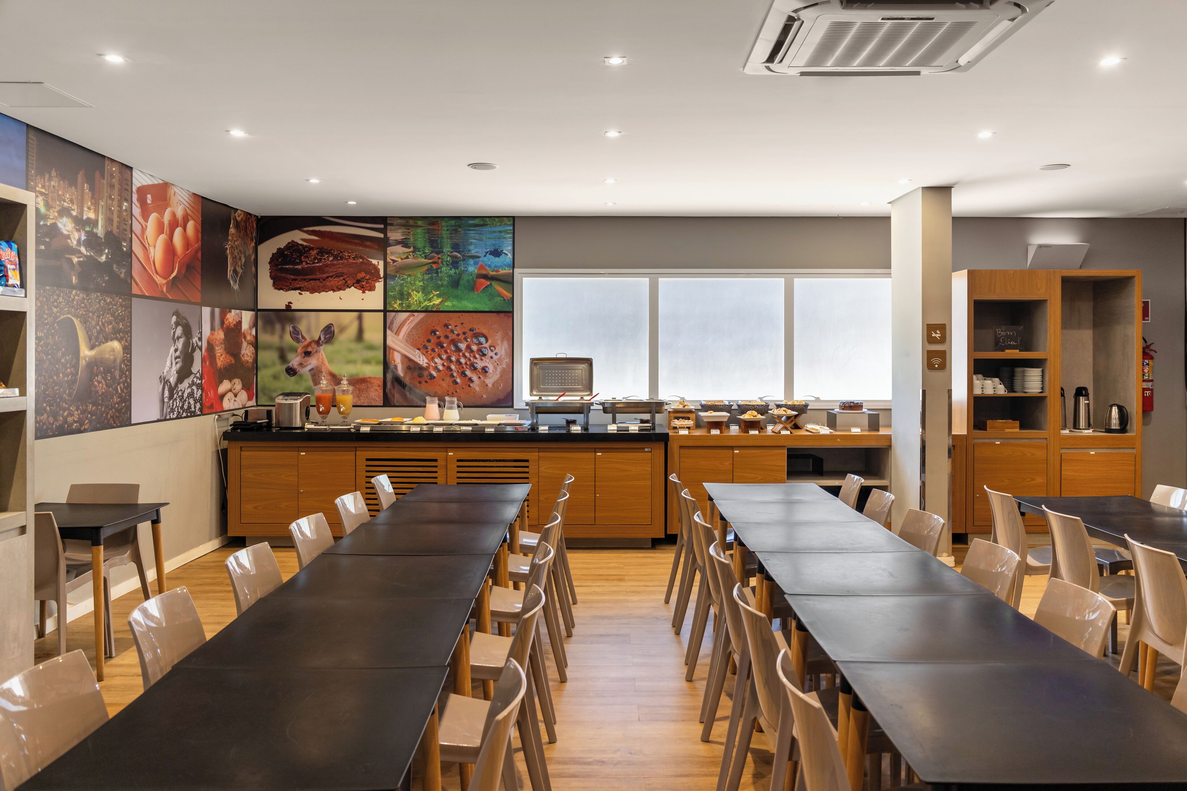 A modern dining area with long tables and chairs, a kitchen counter with various food items, and a large window providing natural light.