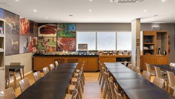 Un comedor moderno con mesas y sillas largas, una encimera de cocina con diversos alimentos y un amplio ventanal que proporciona luz natural.