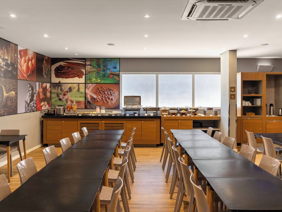 Un comedor moderno con mesas y sillas largas, una encimera de cocina con diversos alimentos y un amplio ventanal que proporciona luz natural.