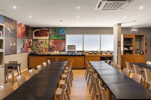 Uma área de refeições moderna com mesas longas e cadeiras, um balcão de cozinha com diversos alimentos, e uma janela grande que proporciona luz natural.