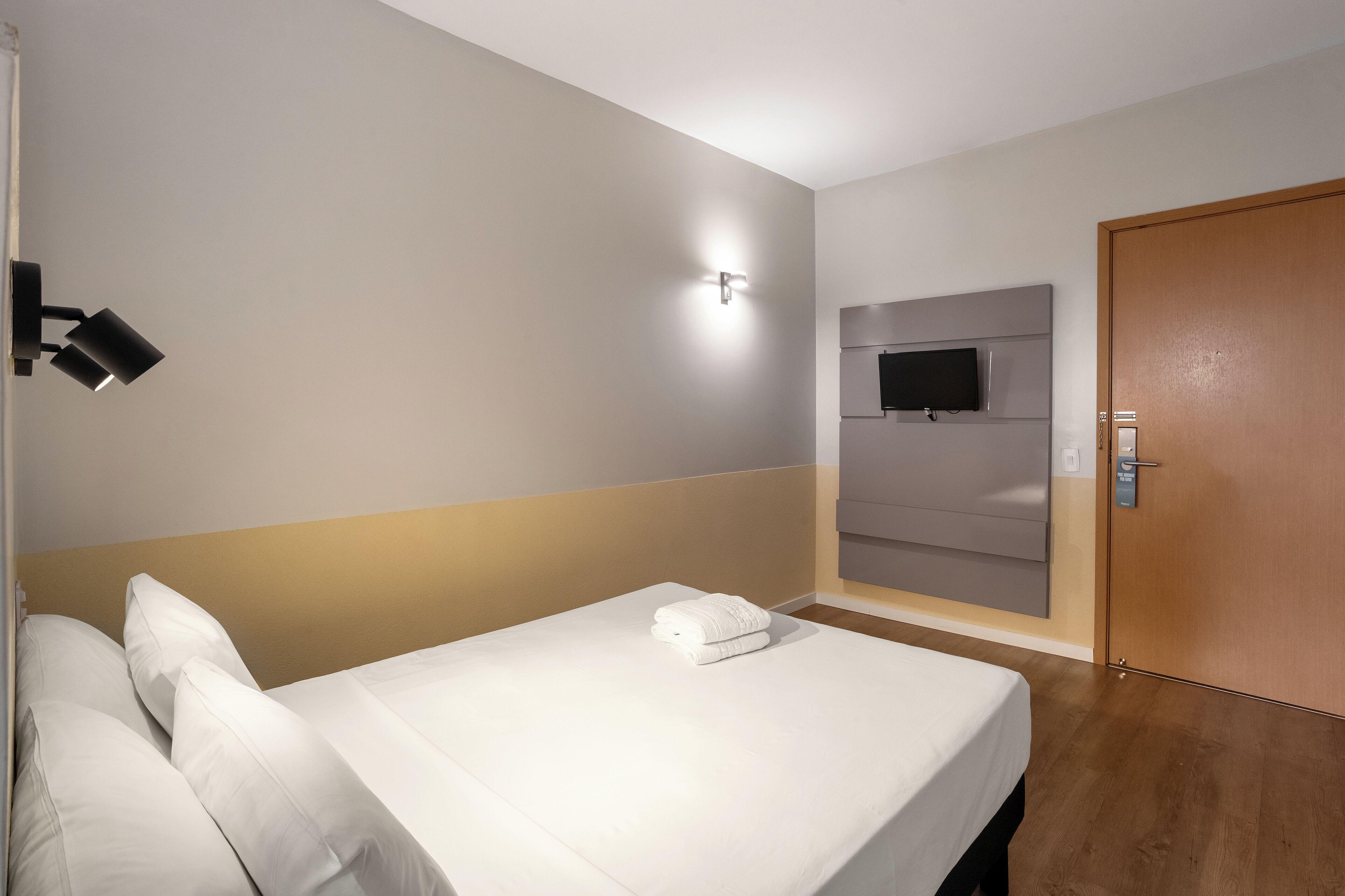 Une chambre d’hôtel avec un lit soigneusement fait, un téléviseur à écran plat fixé au mur et une porte en bois.