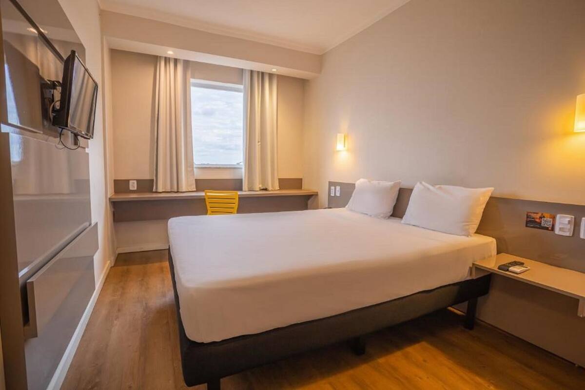 A hotel room with a bed, a TV, a window with curtains, a bedside table, and a wall-mounted light.