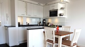A modern kitchen with white cabinetry, a black countertop, and a wooden dining table with white chairs. - Die kleine Residenz (Sylt)