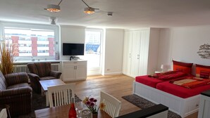 A modern, well-lit living room with a sofa, a dining table, a TV, and a bed with red bedding. - Die kleine Residenz (Sylt)