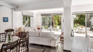 A modern living room with a sofa, dining table, and chairs. Large windows provide a view of the outdoors. - Villa Hibiscus is a unique and spectacular four bedroom villa in an residential gated cul de sac (Protaras)