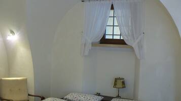 A bedroom with two beds, a chair, a window with white curtains, and a lamp.