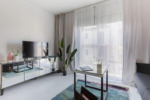 A modern living room with a flat-screen TV on a glass cabinet, a glass coffee table with a magazine, a sofa, and a large window with sheer curtains.
