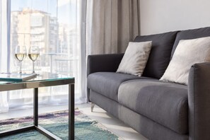 Een moderne woonkamer met een grijze bank, een glazen salontafel en een stadsgezicht door grote ramen.