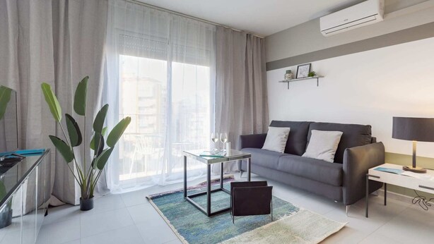 Una sala de estar moderna con un sofá gris, una mesa de centro de vidrio y una silla negra. Hay un ventanal con cortinas translúcidas, una planta en maceta y un equipo de aire acondicionado en la pared.
