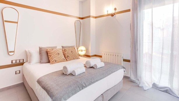 Une chambre d’hôtel avec un grand lit, un miroir, une lampe fixée au mur et une fenêtre avec des rideaux légers.