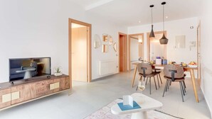 Un salon moderne avec un téléviseur, un coin repas comprenant des chaises et une table, et une cuisine visible à travers une porte ouverte.
