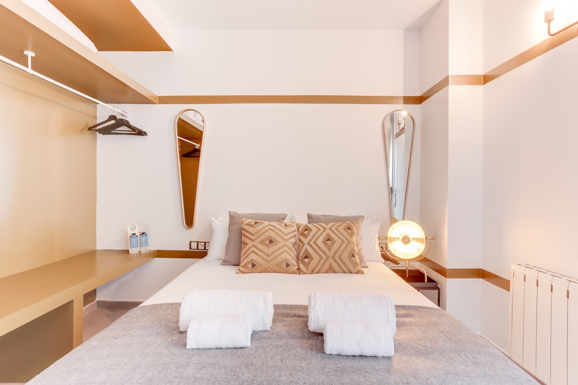 Hotelkamer met een bed, twee handdoeken, een spiegel, een nachtkastje met toiletartikelen en een wandlamp.