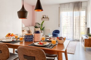Un comedor con una mesa de madera dispuesta para el desayuno, incluyendo platos de fruta, tortitas y zumo de naranja. Hay dos lámparas colgantes sobre la mesa.