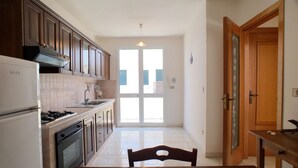 A kitchen with a stove, oven, and sink, leading to a door with a view of buildings outside. - Appartamento Acton - Piano Terra (Ugento)