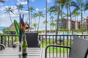 Een balkon met rieten meubilair, een fles Martiniello Sprankelende Cider, en een tropisch uitzicht met palmbomen en een zwembad.