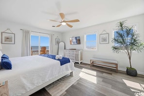 A bedroom with a bed, ceiling fan, windows, a bench, a dresser, and a potted plant. - Salt Therapy OBX | Oceanfront | Dog Friendly, Hot Tub | Nags Head (Nags Head)