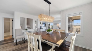 A modern dining area with a large wooden table, white chairs, and a hanging light fixture. - Salt Therapy OBX | Oceanfront | Dog Friendly, Hot Tub | Nags Head (Nags Head)