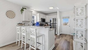 A modern kitchen with white cabinetry, a central island with bar stools, stainless steel appliances, and a view to the outside. - Salt Therapy OBX | Oceanfront | Dog Friendly, Hot Tub | Nags Head (Nags Head)