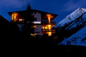 A multi-story building with illuminated windows and balconies, surrounded by snow-covered mountains. - Fernerblick Apartments (Tux)