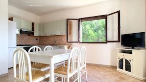 Une cuisine compacte avec des meubles blancs, une table à manger avec des chaises, une fenêtre donnant sur des arbres et un téléviseur posé sur un meuble.