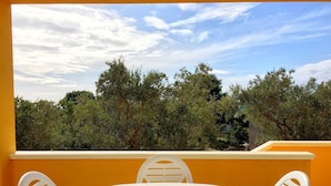 Un balcon avec une table et des chaises blanches, donnant sur un jardin arboré et un ciel dégagé.