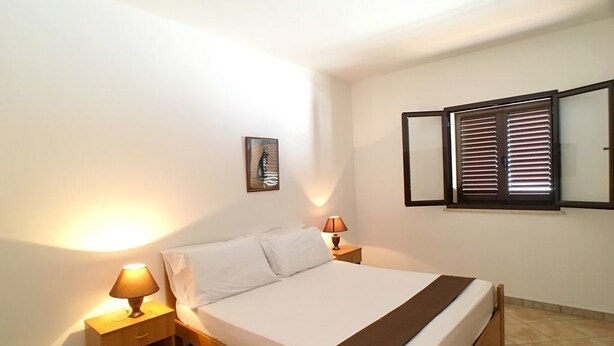 Une chambre à coucher avec un lit, deux lampes de chevet, une table de chevet, un tableau encadré au mur et une fenêtre avec des stores.